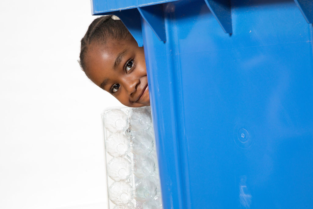 Girl hiding behind blue recycling bin. 